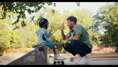 Un-Niño-Pequeño-Con-Un-Suéter-Azul-Y-Un-Casco-De-Seguridad-Negro-Y-Su-Padre-Con-Cabello-Rizado-Y-Una-Camiseta-Verde-Se-Saludan-En-Una-Patineta-En-El-Parque.