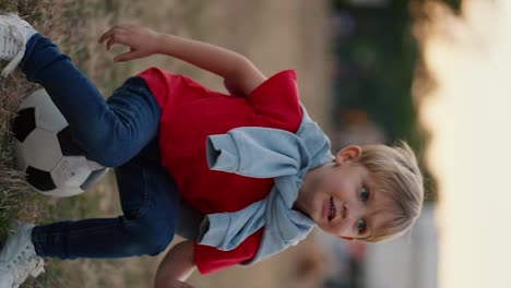 Vídeo-Vertical:-Un-Niño-Rubio-De-Ojos-Azules-Con-Una-Camiseta-Roja-Y-Pantalones-Vaqueros-Salta-Sobre-Una-Pelota-De-Fútbol-En-El-Patio-De-Recreo-Del-Parque.-Un-Niño-Pequeño-Se-Divierte-Y-Se-Sienta-En-Una-Pelota-De-Fútbol-En-El-Campo-De-Fútbol-Del-Parque.