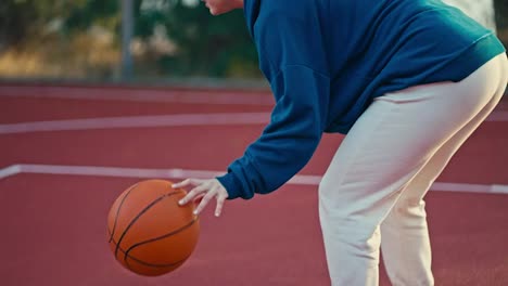 Nahaufnahme-Eines-Blonden-Mädchens-In-Einem-Blauen-Kapuzenpullover-Und-Weißen-Hosen,-Das-Morgens-Während-Ihres-Trainings-Auf-Dem-Red-Street-Court-Einen-Orangefarbenen-Basketball-Schlägt