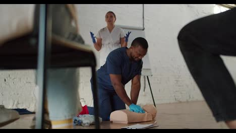 Bottom-view:-A-Black-male-doctor-performs-artificial-respiration-on-a-medical-mannequin,-and-his-assistant-girl-nurse-explains-to-the-public-against-a-white-brick-wall