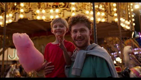 Portrait-of-a-happy-blond-boy-in-a-red-T-shirt-and-his-dad-eating-pink-cotton-candy-in-the-amusement-park-opposite-against-the-backdrop-of-a-beautiful-bright-attraction-in-yellow-colors-in-the-evening