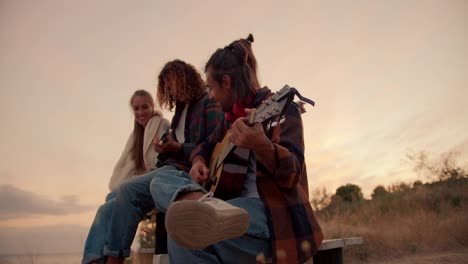 Dos-Chicos-Están-Sentados-Y-Tocan-La-Guitarra.-Dos-Chicas-Escuchan-A-Sus-Novios-Tocar-La-Guitarra-Con-El-Atardecer-De-Fondo.-Descanso-Fuera-De-La-Ciudad.-Descanso-En-La-Casa-De-Campo.