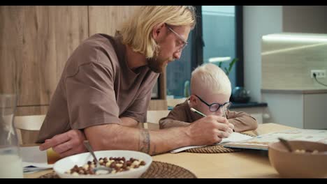 Ein-Blonder-Mann-Mit-Bart-Und-Brille-Hält-Einen-Grünen-Bleistift-In-Den-Händen-Und-Zeigt-Seinem-Kleinen-Albino-Sohn-Mit-Blauer-Brille,-Was-Er-Für-Seine-Hausaufgaben-Machen-Muss,-Während-Er-Sich-In-Der-Küche-Einer-Modernen-Wohnung-Auf-Die-Schule-Vorbereitet