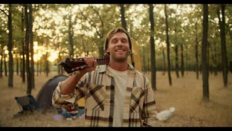 Portrait-of-a-happy-blond-man-with-stubble-in-a-checkered-shirt-who-holds-a-guitar-on-his-shoulder-while-resting-and-taking-a-break-during-a-hike-in-a-sunny-summer-green-forest