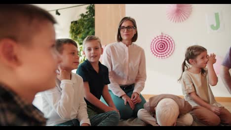 Una-Chica-Rubia-Con-Un-Peinado-Bob-Con-Gafas-Y-Una-Camisa-Blanca-Y-Un-Hombre-Con-Una-Camisa-Violeta-Están-Sentados-En-Un-Círculo-Con-Niños-En-El-Suelo-Y-Hablando-En-Su-Lección-Introductoria-Sobre-Cómo-Prepararse-Para-La-Escuela.