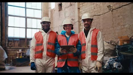 Portrait-of-a-happy-multiracial-trio-in-special-white-uniforms-and-orange-vest-employees-stand-near-a-large-pile-of-garbage-at-a-large-waste-processing-plant