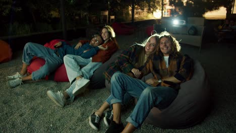 Two-couples-in-love-are-sitting-on-beanbag-chairs-and-watching-a-movie.-A-party-outside-the-city.-Rest-in-the-country-house-at-the-weekend