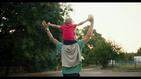 Rear-view-of-a-little-blond-boy-sitting-on-his-dad's-shoulders-in-a-Green-T-shirt,-they-hold-hands-and-raise-them-up.-Happy-little-boy-sitting-on-the-shoulders-of-his-dad-who-is-walking-in-the-park