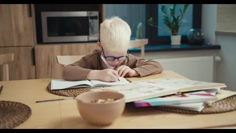 Un-Pequeño-Niño-Albino-Con-Gafas-Azules-Escribe-Algo-En-Su-Cuaderno-Y-Se-Prepara-Para-El-Día-Siguiente-En-La-Escuela-En-Casa-En-La-Cocina-En-Un-Apartamento-Moderno-Por-La-Noche