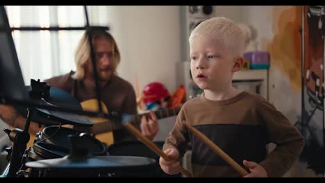 A-happy-albino-boy-with-white-hair-and-blue-eyes-plays-on-an-electric-musical-set-using-special-sticks,-and-a-father,-a-blond-man-in-glasses-with-a-beard,-helps-his-son-and-plays-along-with-him-on-the-guitar