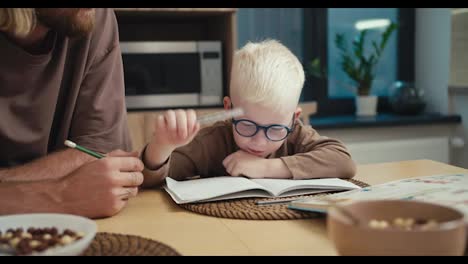 Nahaufnahme-Eines-Kleinen-Albinojungen-Mit-Weißem-Haar-Und-Blauer-Brille,-Der-Seinen-Stift-Hinlegt-Und-Seine-Beiden-Handflächen-In-Die-Höhe-Hebt,-Um-Bei-Seinen-Mathehausaufgaben-An-Seinen-Fingern-Zu-Zählen