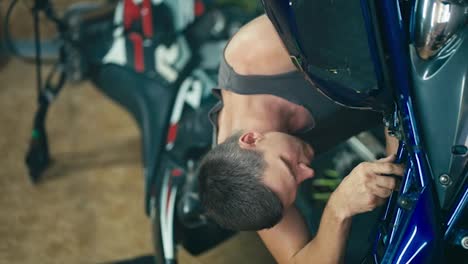 Vertical-video-of-a-man-with-a-short-haircut-in-a-gray-T-shirt-repairing-his-blue-bike-with-a-screwdriver-against-the-background-of-another-motorcycle-in-his-garage-workshop