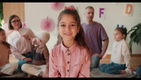 Ein-Glückliches,-Fröhliches-Mädchen-Mit-Braunen-Augen-In-Einem-Rosa-Hemd-Blickt-In-Die-Kamera-Und-Lächelt-Vor-Dem-Hintergrund-Ihrer-Ersten-Schulvorbereitungsstunde-Mit-Ihren-Lehrern-Und-Einer-Gruppe-Von-Kindern