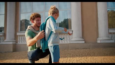 Un-Padre-Con-El-Pelo-Rizado-Y-Una-Camiseta-Verde-Envía-A-Su-Pequeño-Hijo-Rubio-Con-Una-Chaqueta-Azul-Y-Una-Mochila-Turquesa-A-La-Escuela.-Un-Pequeño-Estudiante-De-Primaria-Corre-A-La-Escuela.