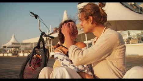 Happy-couple-on-a-date,-a-blond-guy-in-light-clothes-straightens-the-hair-of-a-brown-haired-girl-in-light-clothes-with-a-black-bow-who-sits-near-a-bicycle-on-the-beach