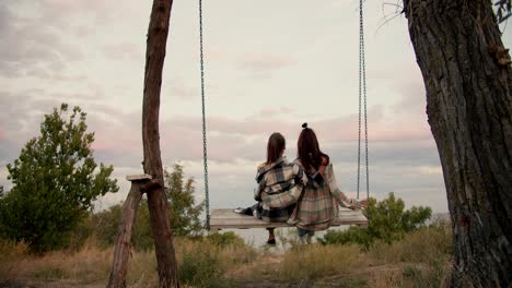 Close-up-shooting-from-behind:-A-brunette-girl-in-a-checkered-shirt-and-a-blonde-girl-are-swinging-on-a-wooden-swing-outside-the-city.-Rest-in-the-country-by-the-sea