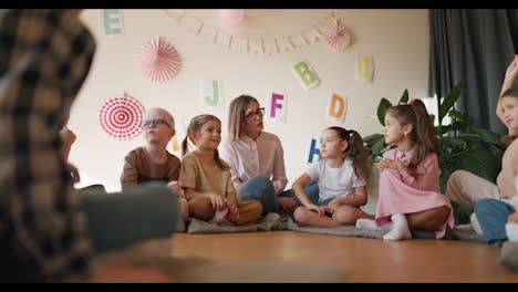 Una-Niña-Con-Un-Peinado-Bob,-Gafas-Y-Una-Camisa-Blanca-Se-Comunica-Con-Sus-Estudiantes-Mientras-Está-Sentada-En-El-Suelo-Sobre-Una-Almohada-Especial-En-Un-Club-Para-Preparar-A-Los-Niños-Para-La-Escuela.