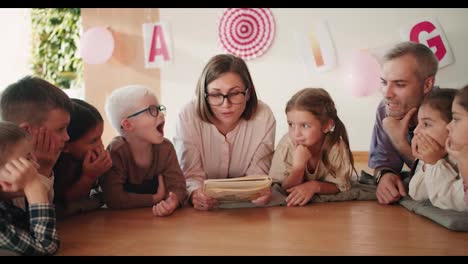 Eine-Lehrerin-Mit-Bob-Frisur,-Brille-Und-Weißem-Hemd-Liegt-Mit-Den-Kindern-Auf-Speziellen-Kissen-Auf-Dem-Boden-Und-Liest-Ihnen-In-Ihrer-Ersten-Unterrichtsstunde-Zur-Vorbereitung-Auf-Die-Schule-Im-Schulvorbereitungsclub-Der-Kinder-Ein-Buch-Vor