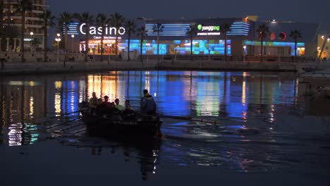 Vista-Nocturna-Del-Paseo-Marítimo-De-La-Ciudad-Y-Bote-De-Remos-Navegando-En-El-Puerto