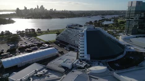 Toma-Aérea-Acercándose-Al-Lujoso-Hotel-Crown-Casino-Con-El-Río-Swan-Y-El-Horizonte-De-La-Ciudad-De-Perth-Al-Fondo---Hora-Del-Atardecer-En-Australia-Occidental