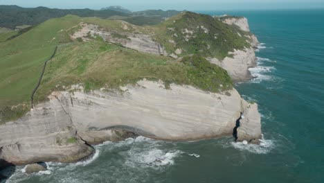 Stunning-aerial-view-rising-above-the-remote-wilderness-coastal-landscape-of-Cape-Farewell-headland-with-rough-and-wild-ocean-waves-breaking-against-cliffs-in-New-Zealand-Aotearoa