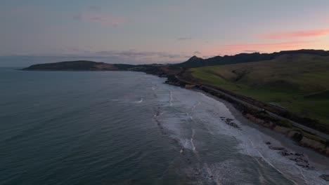 Scenic-aerial-view-of-ocean-with-gentle-rolling-waves-during-sunset-with-orange-glow-in-sky-at-Katiki-peninsula-in-North-Otago,-New-Zealand-Aotearoa