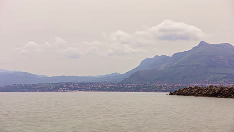 Timelapse-shot-of-mountain-range-along-the-seashore-with-town-houses-in-the-foreground-in-Sicily,-Italy-on-a-cloudy-day