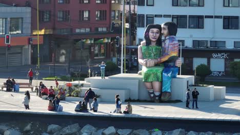 Families-and-people-walking,-taking-photos,-and-enjoying-the-sunny-day-around-the-sculpture-‘Sentados-frente-al-mar’-in-Puerto-Montt,-Chile