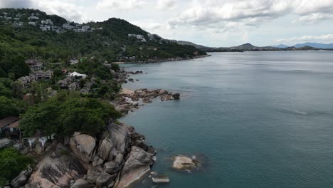 Left-to-right-pan-shot-of-a-rocky-coast-with-green-hills-and-clear-ocean-water-Koh-Samui-Thailand