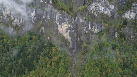 Left-to-right-aerial-shot-of-a-thin-stream-waterfall-flowing-from-a-high-mountain-summit-with-clouds-and-mist-all-around-the-place-Klöntalersee-Glarus-Switzerland