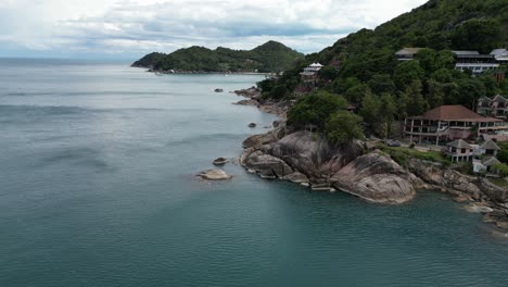 Panoramic-aerial-shot-of-rocky-coast-with-visible-resort-at-the-beach-with-lots-of-trees-and-green-vegetation-trees-Koh-Samui-Thailand