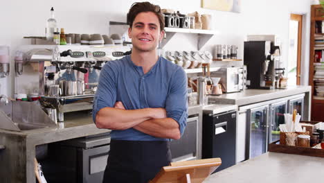 Young-male-coffee-shop-owner-crossing-arms-behind-counter