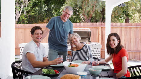 Parents-and-adult-children-at-table-in-garden-look-to-camera