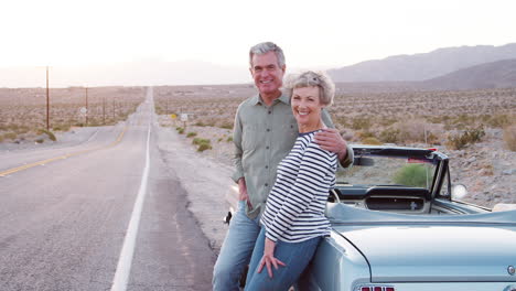 Senior-couple-on-road-trip-standing-by-car-smiling-to-camera