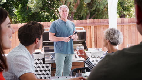 Family-at-a-table-outdoors-turn-to-dad-standing-by-barbecue