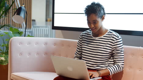 Close-up-of-mixed-race-businesswoman-working-on-a-laptop-computer-sitting-on-a-sofa-in-a-casual-office-smiling