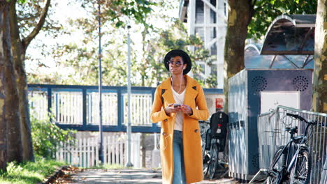 Fashionable-young-black-woman-wearing-a-hat,-sunglasses,-an-unbuttoned-yellow-pea-coat-and-jeans-walking-along-a-treelined-street-looking-around-and-using-smartphone,-low-angle