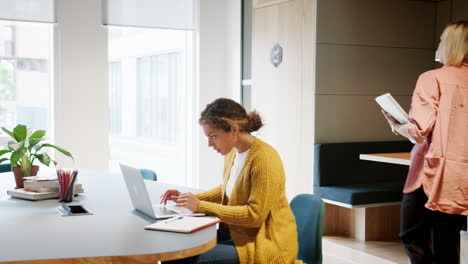 Young-adult-woman-working-at-laptop-computer-sitting-in-an-office-while-her-colleague-enters-the-room-and-sits-down-to-work,-handheld