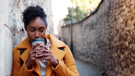 Millennial-black-woman-wearing-a-yellow-coat-leaning-on-a-stone-wall-in-an-alleyway-drinking-a-takeaway-coffee,-focus-on-foreground