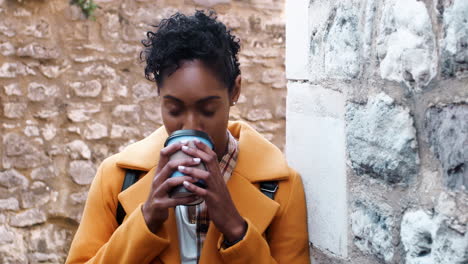 Millennial-black-woman-wearing-a-yellow-coat-leaning-on-a-stone-wall-in-an-alleyway-drinking-a-takeaway-coffee,-close-up