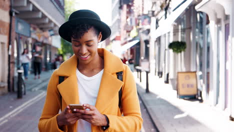 Millennial-black-woman-wearing-a-yellow-pea-coat-and-a-homburg-hat-using-her-smartphone,-standing-in-the-street-on-a-sunny-day,-close-up