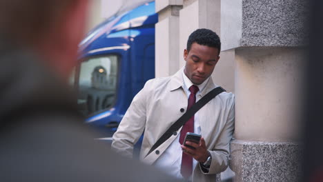Young-black-businessman-leaning-on-a-wall-in-the-street-in-London-using-his-smartphone,-selective-focus,-waist-up