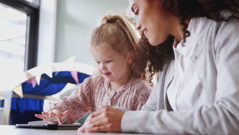 Female-infant-school-teacher-working-with-a-young-white-schoolgirl-using-tablet,-close-up,-low-angle