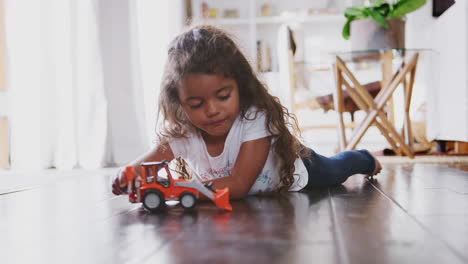 Young-Hispanic-girl-lying-on-the-floor-in-the-sitting-room-playing-with-toy-digger-truck,-side-view