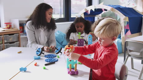 Female-infant-school-teacher-sitting-at-table-with-children-using-construction-toys,-lens-flare