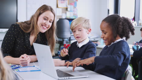 Female-school-teacher-sits-with-two-kids-using-laptop-computer-in-a-primary-school-class,-close-up