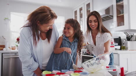 Young-girl-making-cakes-with-her-mum-and-grandmother-filling-cake-forms,-mum-holding-the-mixing-bowl