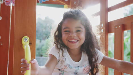 Young-Hispanic-girl-playing-on-a-climbing-frame-in-a-playground-smiling-to-camera,-backlit,-close-up