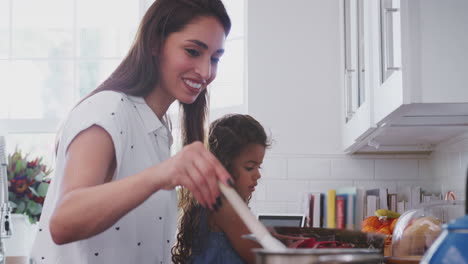 Hispanic-woman-cooking-at-the-hob-in-kitchen-with-her-young-daughter-beside-her,-close-up,-side-view