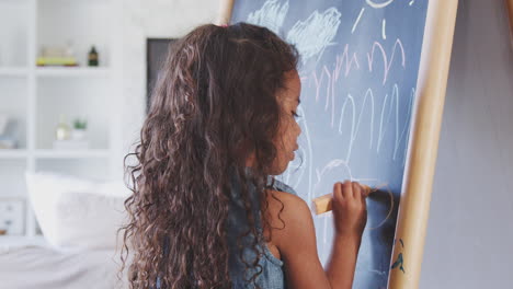 Young-Hispanic-girl-drawing-with-chalks-at-a-blackboard-at-home,-close-up,-back-view